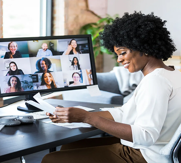 A woman at a desk, holding documents and on a virtual conferencing video call.